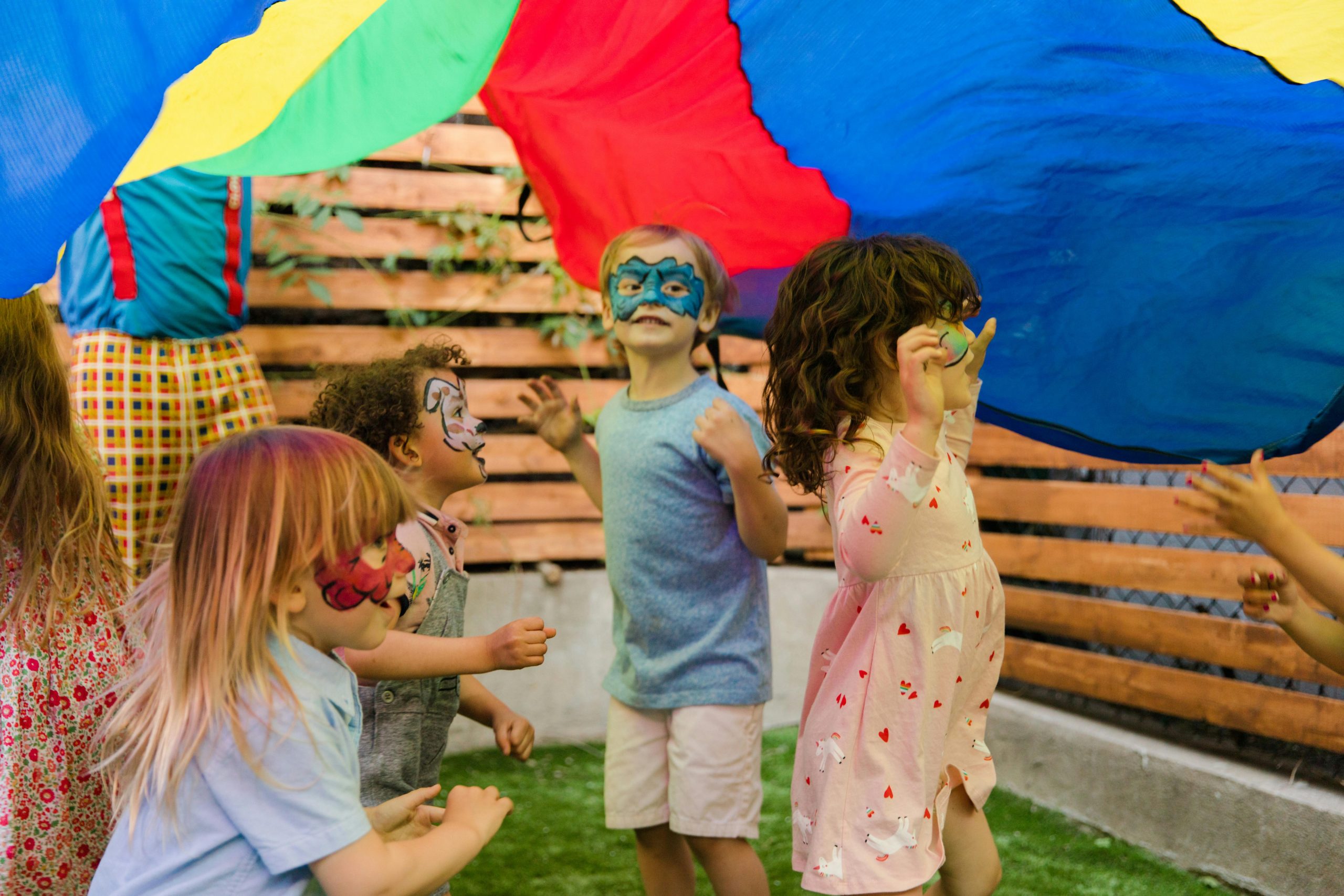 A group of children with their faces painted are playing under a parachute.