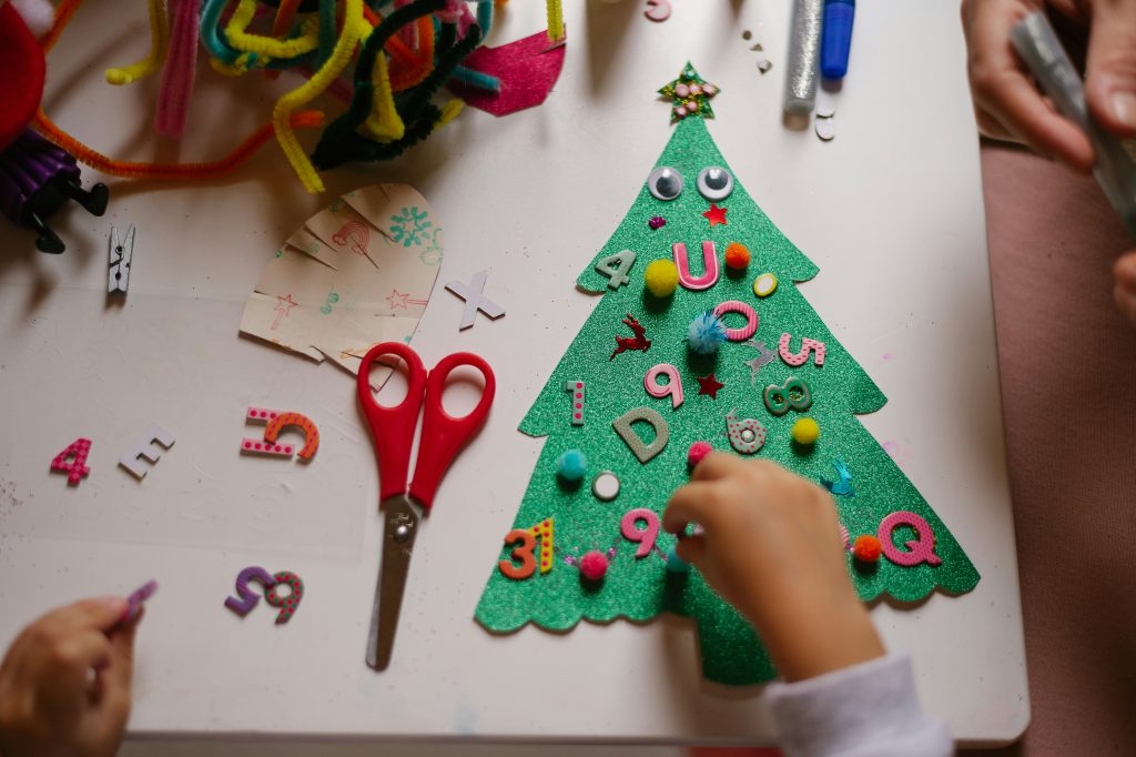 A felt Christmas tree on a white table. We can see a child's hands adding stickers and pompoms to the tree. Beside the tree is a pair of red scissors.