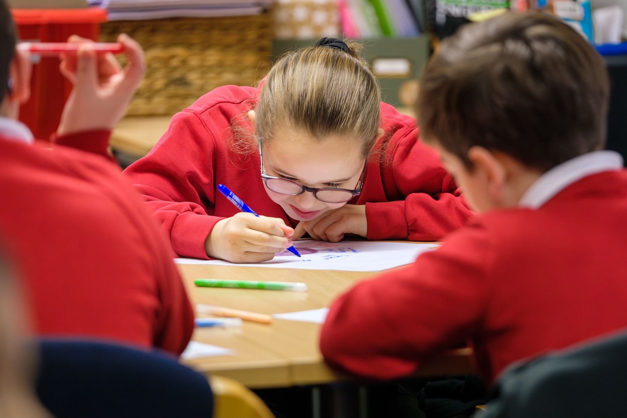 A girl in a red school jumper and glasses concentrates on her craft. In the foreground, we see the backs of two boys in their school jumpers, also working on their craft.