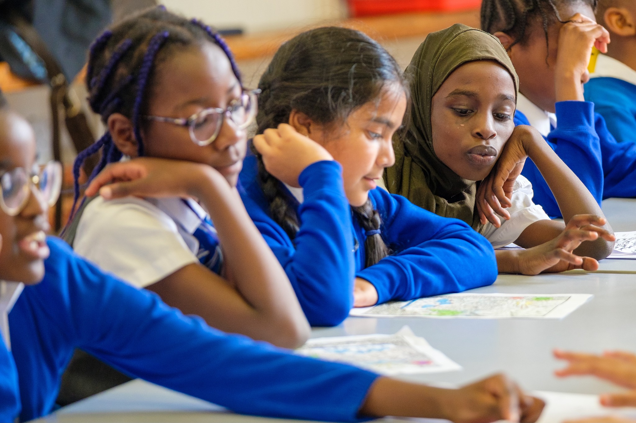 A group of children in school uniform sit at a table. They're working on a colouring activity.