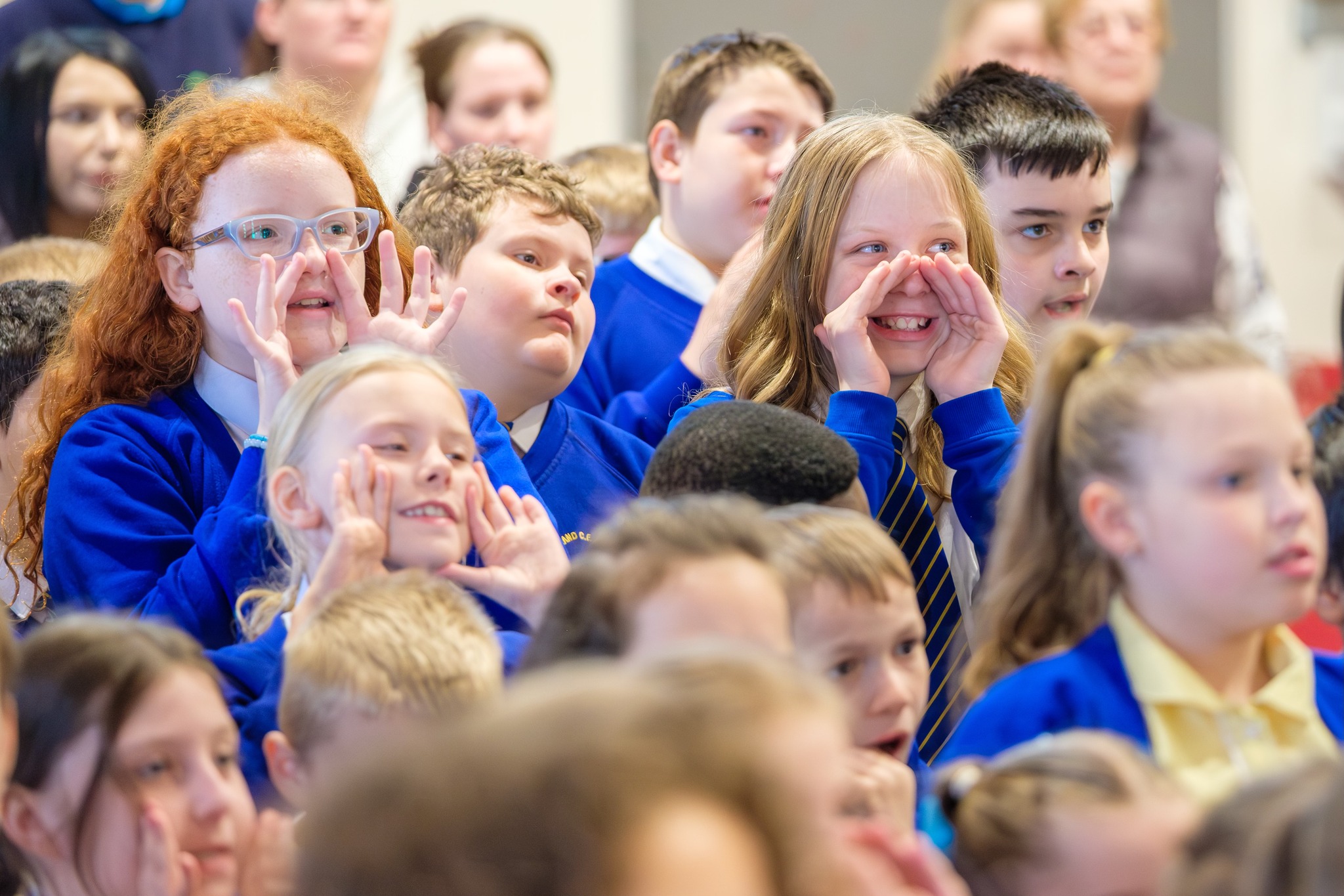 A group of school children in bright blue uniform are standing up and singing. They've got their hands cupped round their mouths and are smiling. In the background, some parents are also taking part.