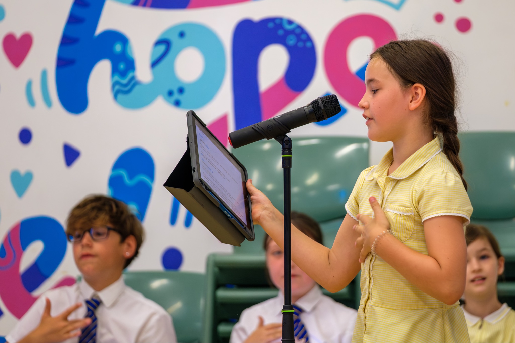 A young girl reads out loud into a microphone from a tablet. Behind her on the wall in colourful letters we can see the word 'hope'.