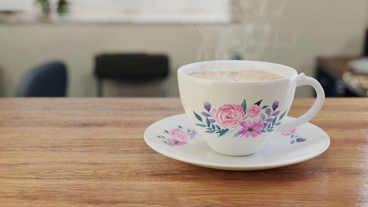 A white floral tea cup full of steam tea sits on a matching saucer. It's placed on a wooden counter top. In the background we can see a window and some plastic chairs.