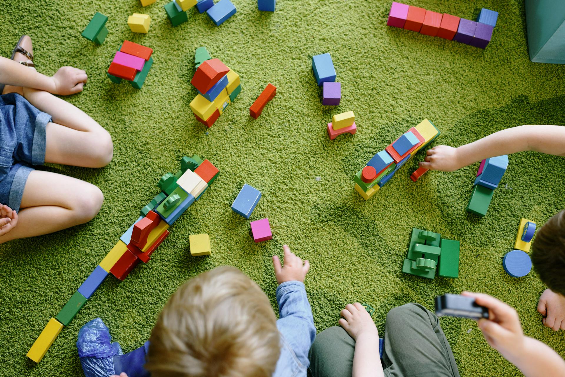 A green mat covered in colourful building blocks. Some young children are building shapes with them.