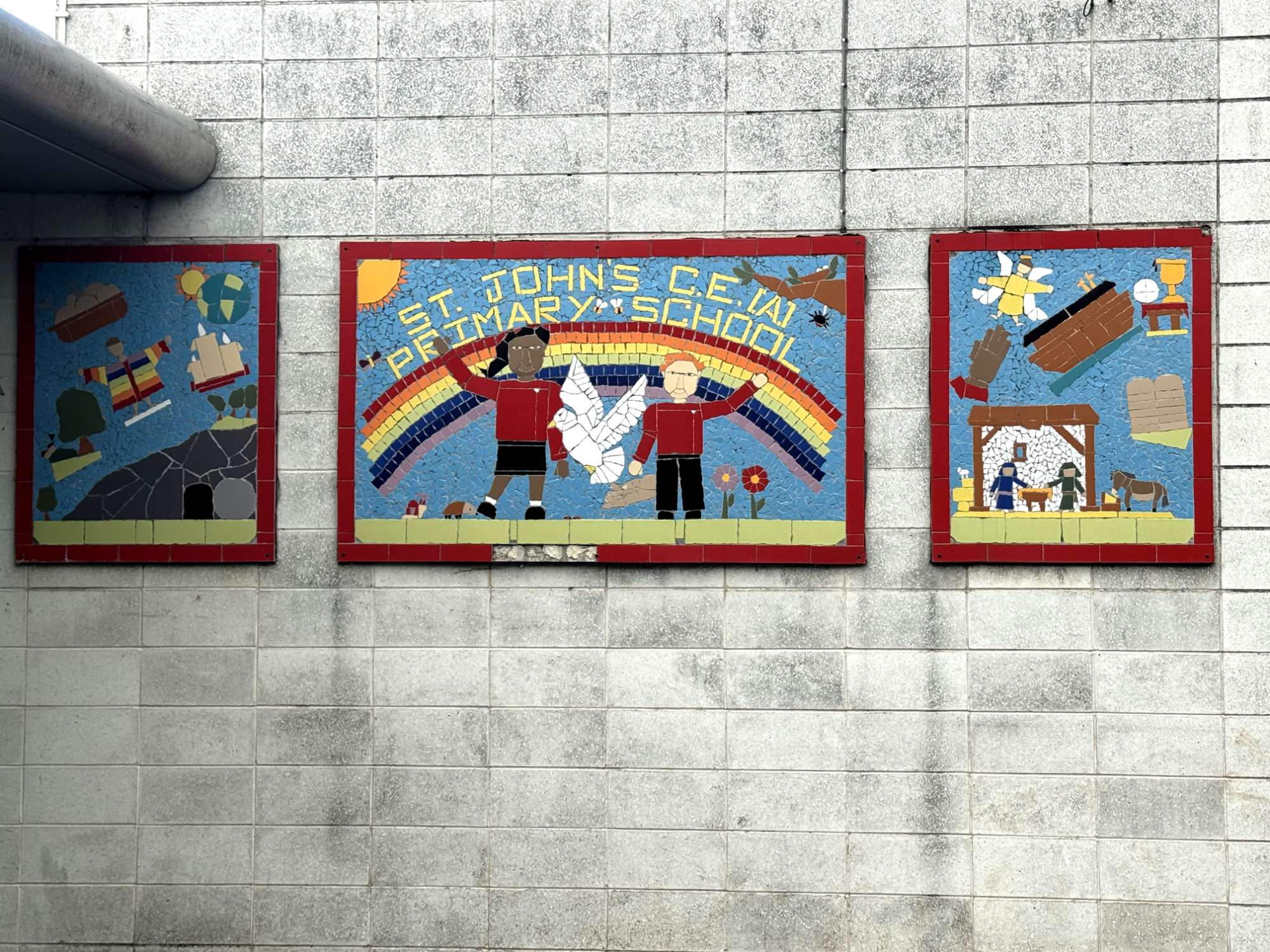 A concrete brick wall. Set into the wall are three colourful mosaics. The central one reads 'St John's CEA Primary School'. It depicts two children in red school jumpers, smiling and waving. In between them is a dove; behind them is a rainbow. The mosaics to the right and left depict stories from the bible: Joseph in a rainbow coat, Jesus' open tomb, the sun and earth, the birth of Jesus, Noah's ark, and so on.
