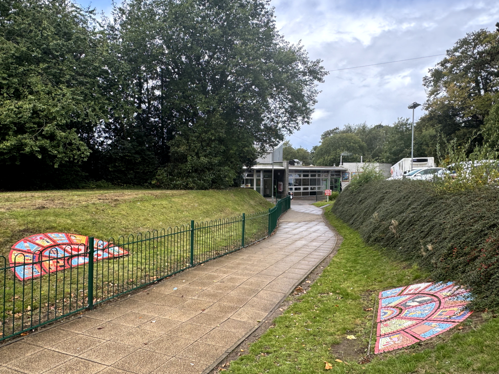 A path, wet with rain, leads between two grass banks to a school building. On either side are two colourful mosaics, set into the grass.